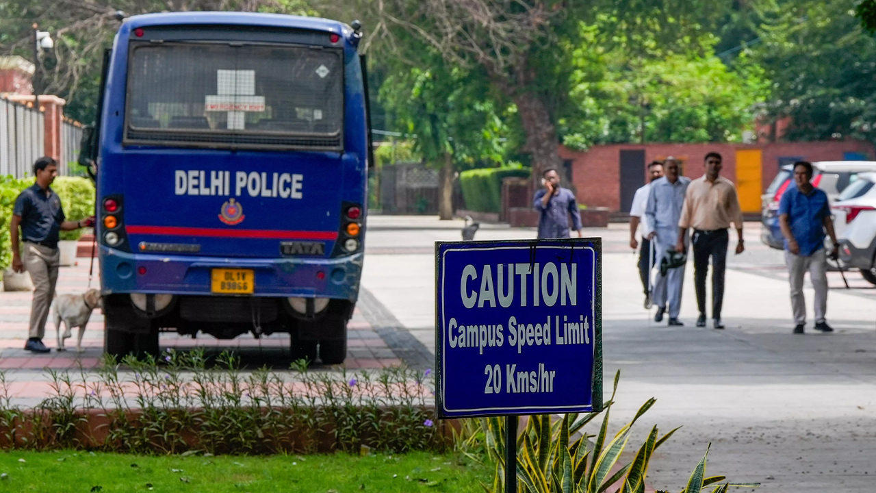Rear view of a blue Delhi Police bus with a security officer and a canine near a campus speed limit sign.